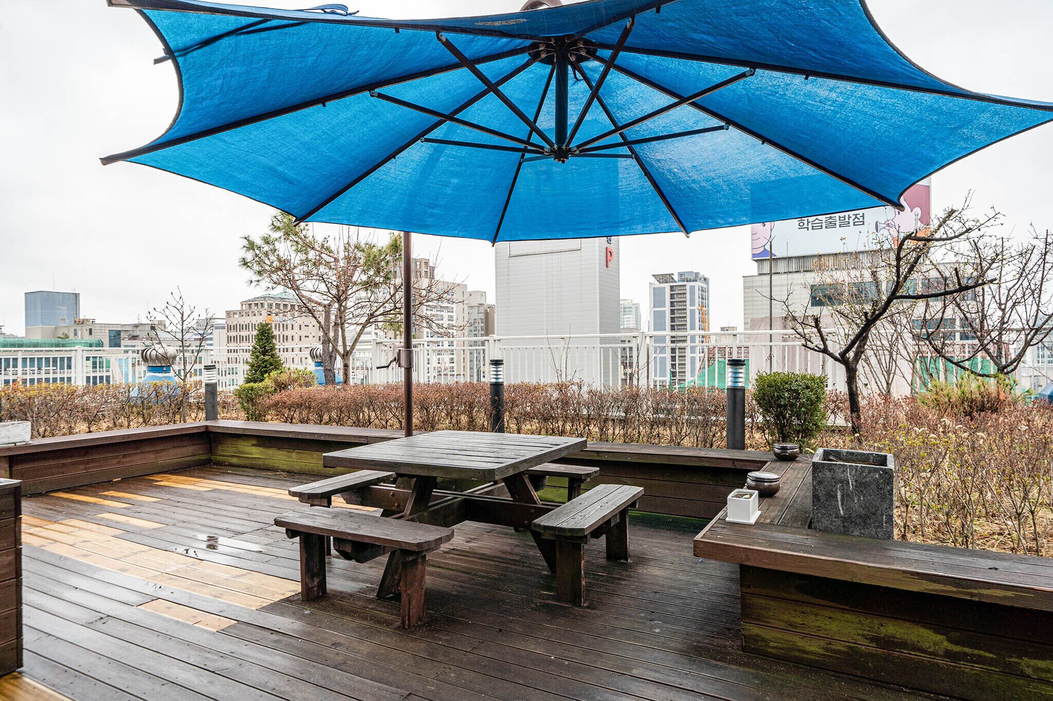 A rooftop patio with wooden flooring, picnic tables, and a large blue umbrella providing shade.