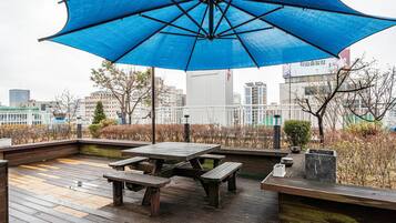 A rooftop patio with wooden flooring, picnic tables, and a large blue umbrella providing shade.
