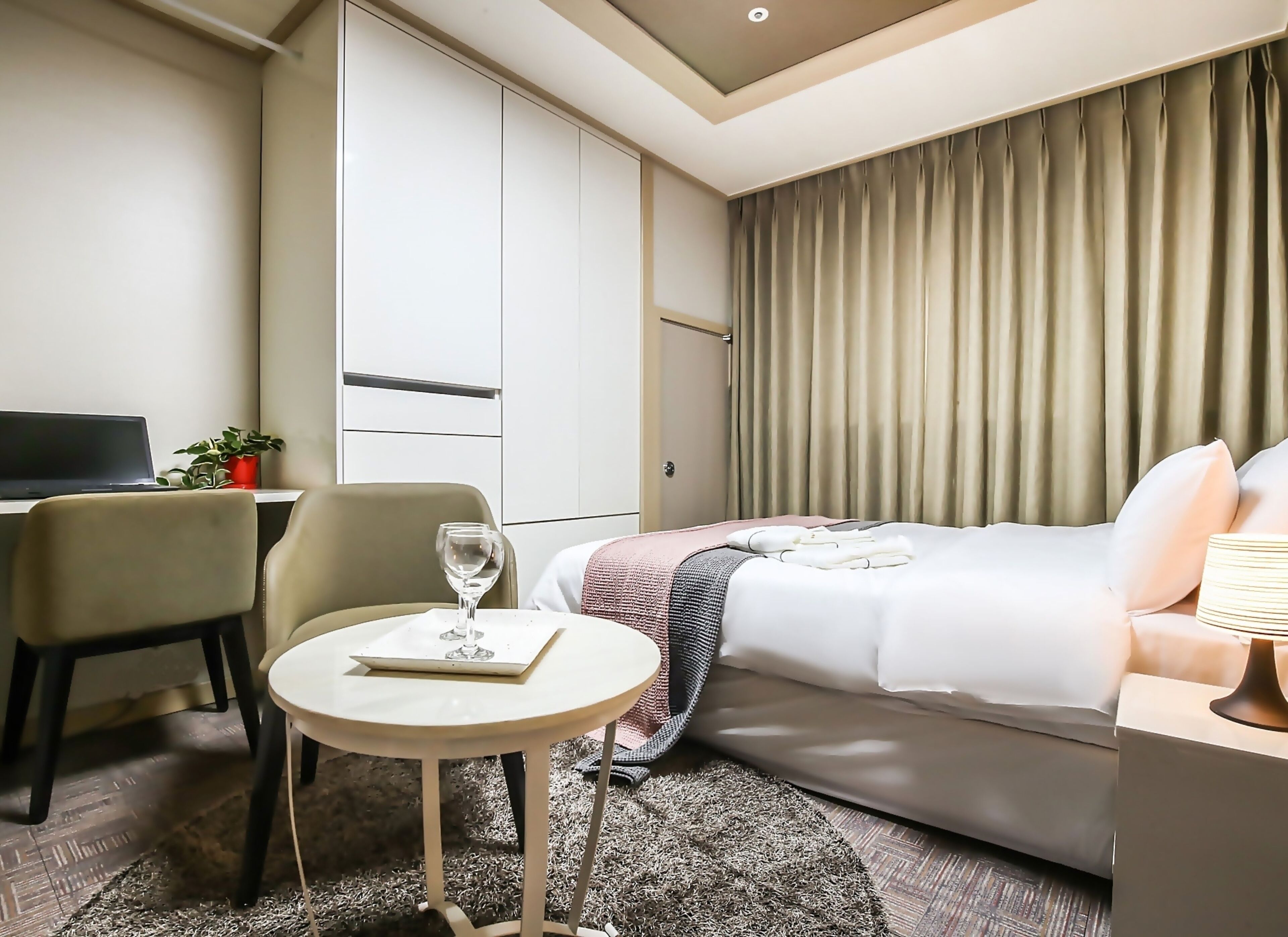 A hotel room with a bed, a desk, a chair, a small table with a glass of water, and a potted plant.