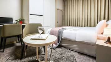 A hotel room with a bed, a desk, a chair, a small table with a glass of water, and a potted plant.