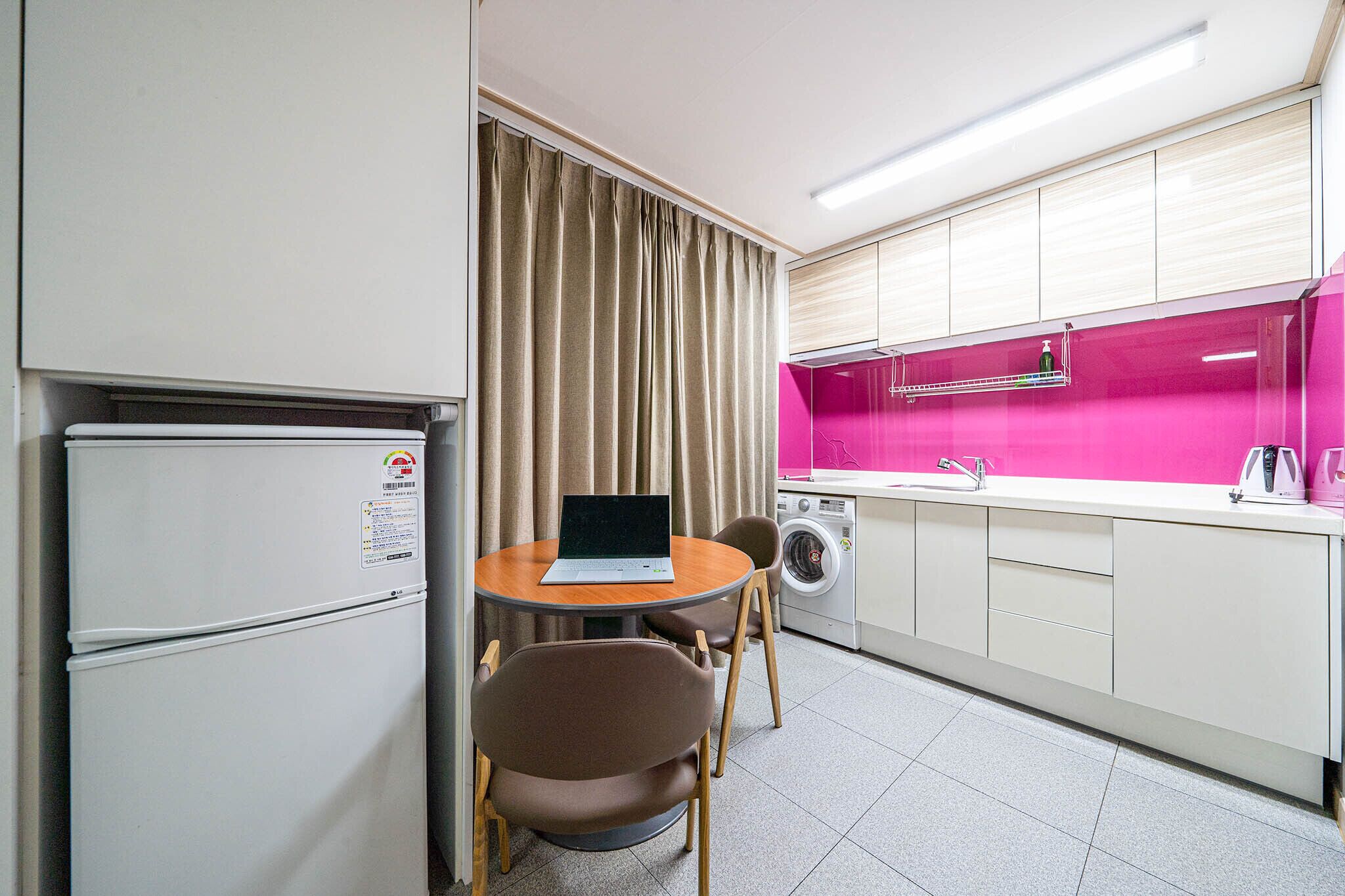 Een compacte kitchenette met een koelkast, wasmachine en een kleine eettafel met stoelen.