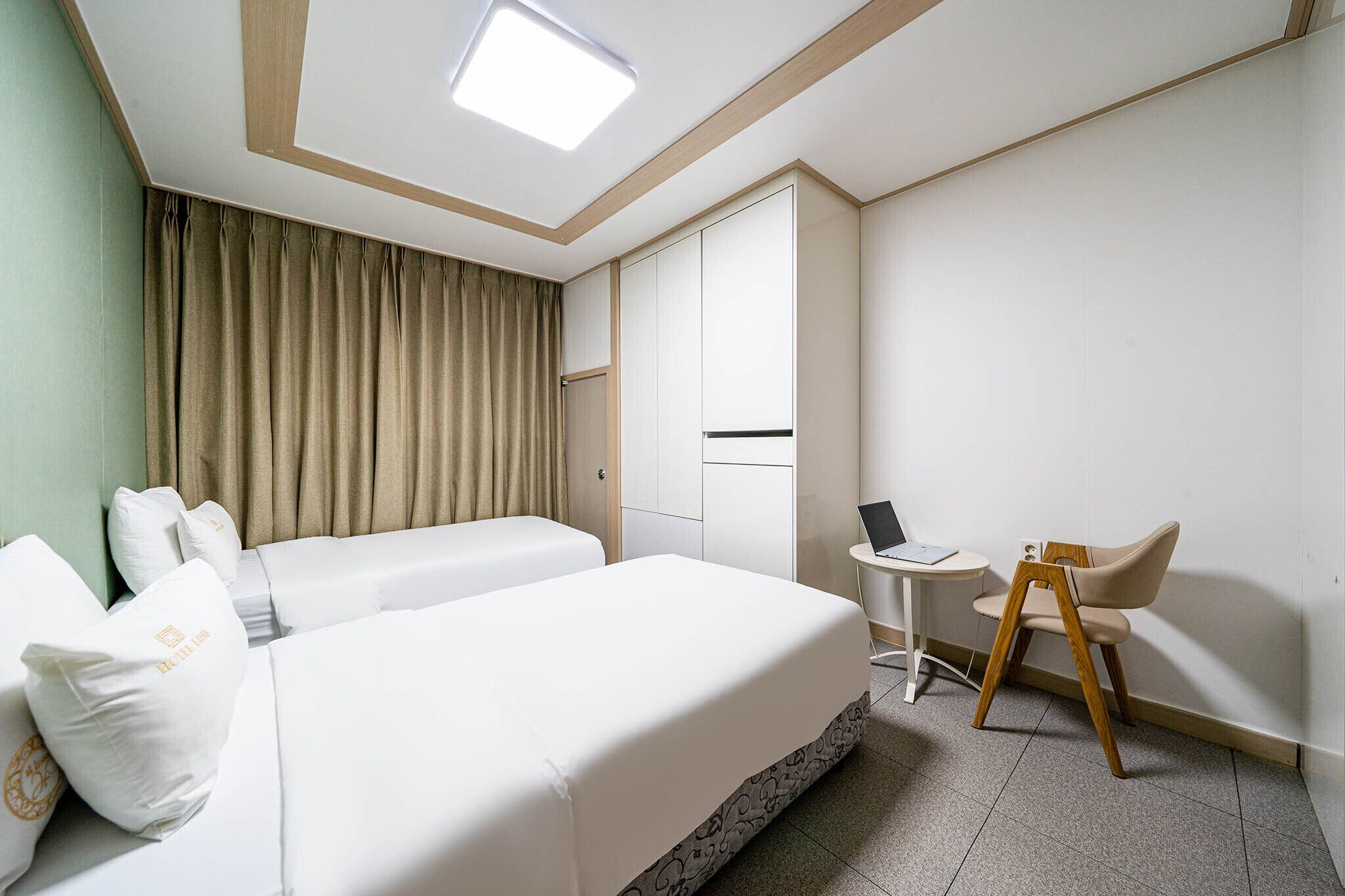 Et hotelværelse med to senge, et skrivebord med en bærbar computer, en stol og et gardin.