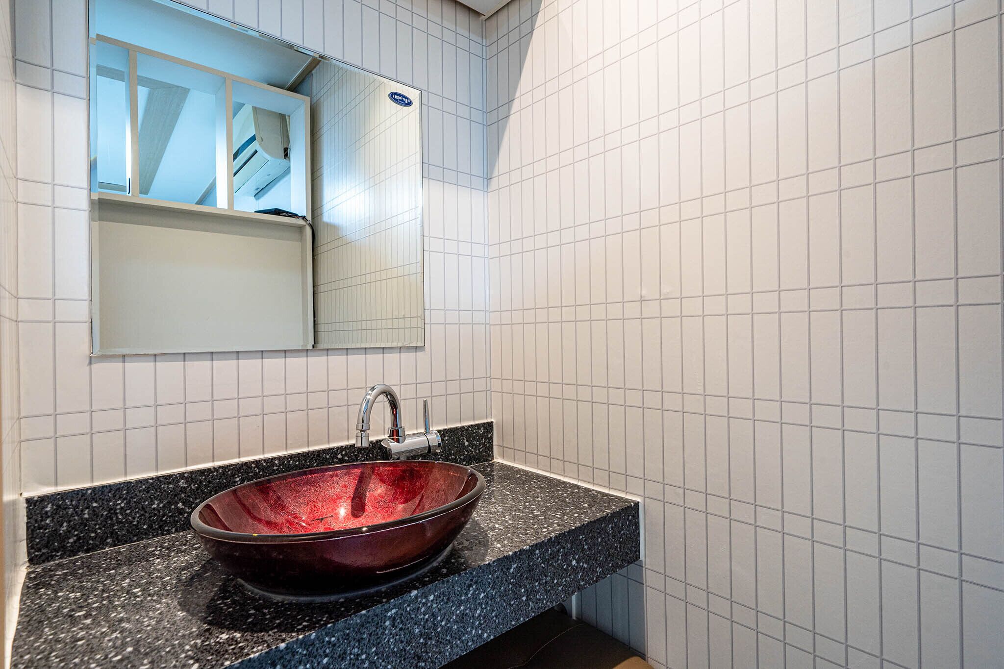 A bathroom with a black granite countertop, a red glass bowl sink, and white tiled walls.