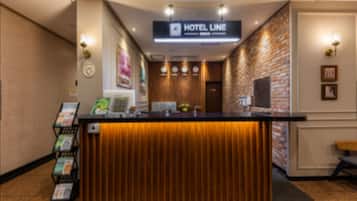 Un mostrador de recepción de hotel con encimera de madera, un letrero que indica la fila de recepción, y un área de espera con pared de ladrillo y una puerta.