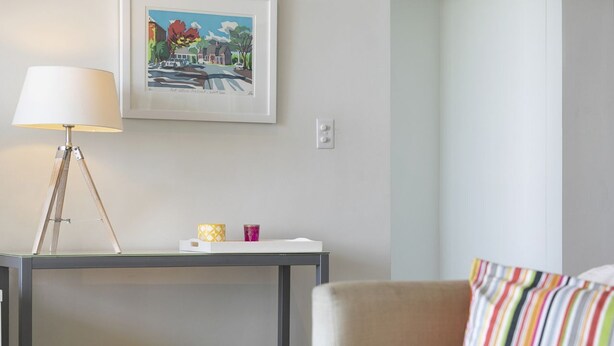 Een kamer met een bed, een nachtkastje met een lamp, een ingelijst schilderij aan de muur en een stoel met een gestreept kussen.
