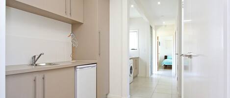 Una cocina moderna con lavavajillas, un lavabo y armarios. Un pasillo conduce a un dormitorio con una cama y una ventana.