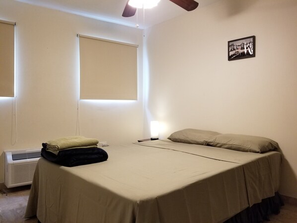 Ein kleines Zimmer mit einem Einzelbett, einer Nachttischlampe, einem Fenster mit Jalousien, einem Wandbild und einem Heizgerät.