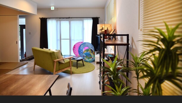 A modern living room with a sofa, a coffee table, and a lamp. There is a window with curtains and a plant in the foreground. - Sun Flower (Onna)