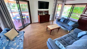 Sala de estar con sofá, una mesa de centro y televisor. Hay una puerta corrediza de vidrio que da a un área exterior con una mesa y sillas.