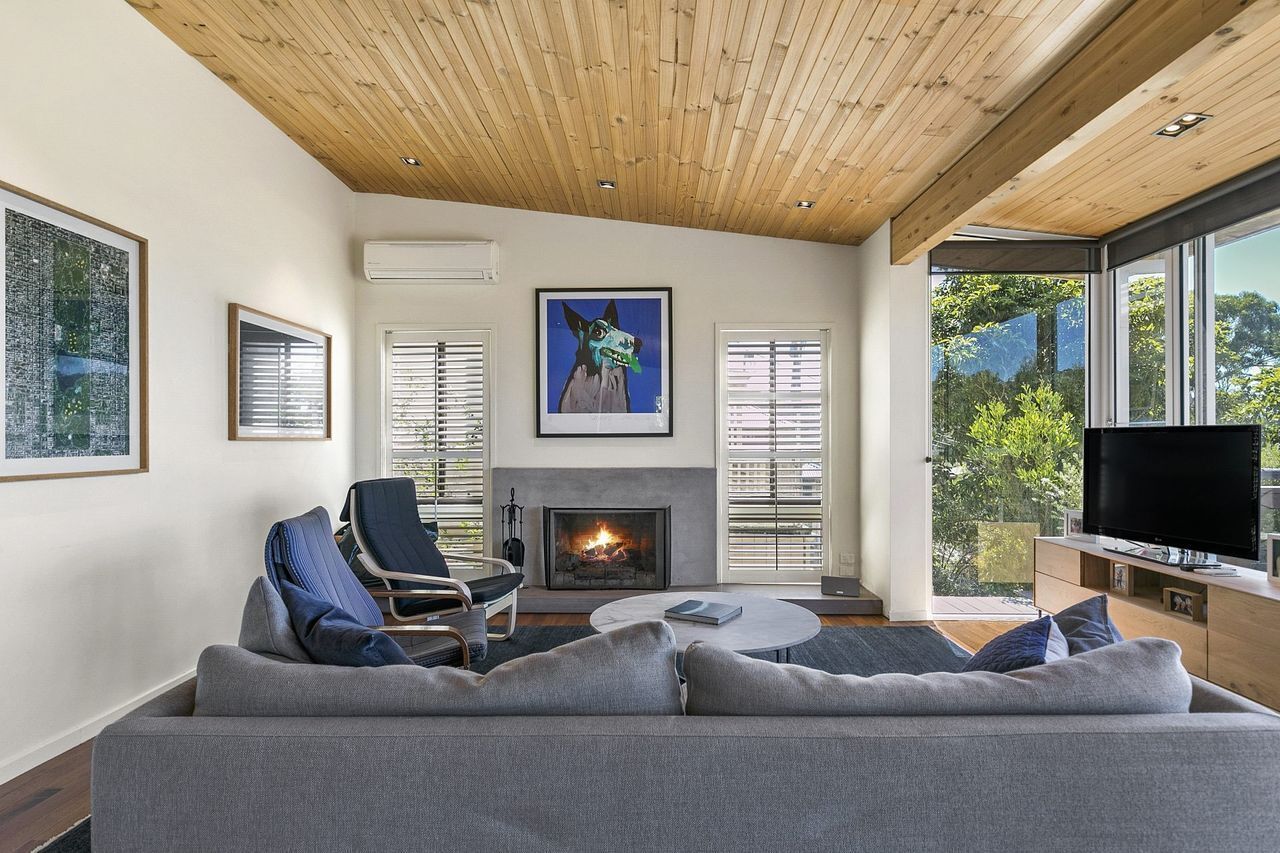 A modern living room with a fireplace, a sofa, a television, and large windows offering a view of the outdoors.