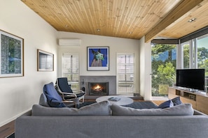 A modern living room with a fireplace, a sofa, a television, and large windows offering a view of the outdoors.
