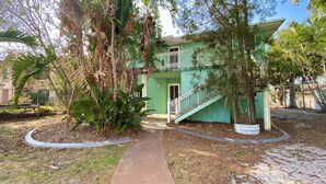 A green building with a staircase leading to a door, surrounded by tropical plants and a paved walkway. - Rio Vista - Burrum Heads - Riverfront - 2BR - Large Yard (BURRUM HEADS)