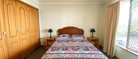 A bedroom with a large wooden wardrobe, a bed with a colorful patterned bedspread, two bedside tables with lamps, and a window with curtains.