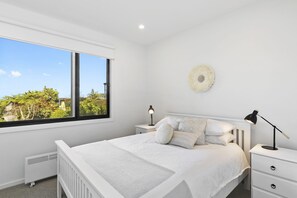 Une chambre avec une grande fenêtre, un lit recouvert d’une literie blanche, deux tables de chevet avec des lampes et un objet décoratif fixé au mur.