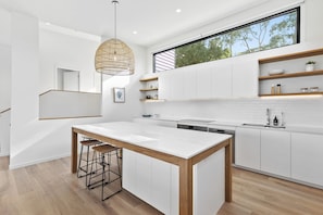 A modern kitchen with a central island, wooden bar stools, white cabinetry, and a large window above the sink. - Belvedere Four (Lorne)