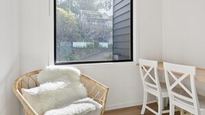 Une pièce d’angle avec une grande fenêtre, un fauteuil en osier avec un coussin blanc et une table blanche avec des chaises.