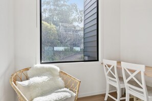 A corner room with a large window, a wicker chair with a white cushion, and a white table with chairs. - Belvedere Four (Lorne)