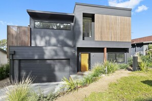 Une maison moderne à deux étages, dotée d’un garage, d’une porte en bois et d’un petit jardin devant, agrémenté de plantes.