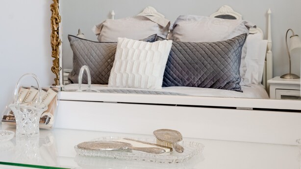 Une chambre à coucher avec un grand miroir décoratif, un lit recouvert de plusieurs coussins, une table de chevet avec une lampe, et un plateau en verre sur lequel reposent un livre et un petit objet décoratif.