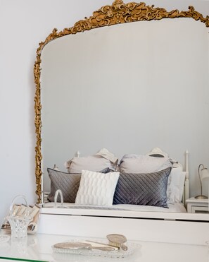Ein Schlafzimmer mit einem großen, verzierten Spiegel, einem Bett mit vielen Kissen, einem Nachttisch mit einer Lampe und einem Glastablett mit einem Buch und einem kleinen Dekorationsgegenstand.
