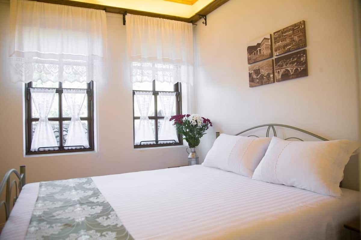 Quarto com cama, duas janelas com cortinas, um vaso com flores e decoração na parede.