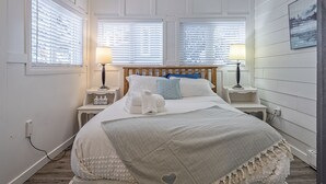 Un dormitorio ordenado con cabecera de madera, dos mesitas de noche con lámparas, cama con ropa de cama blanca y una manta gris, y un cuadro colgado en la pared.
