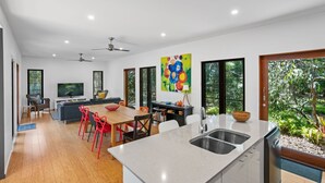 A modern kitchen with a central island, a dining area with red chairs, and a living room with a sofa and TV. - 16 Ibis Court - Rainbow Shores. Wifi. Pool. Quick walk to beach (RAINBOW BEACH)
