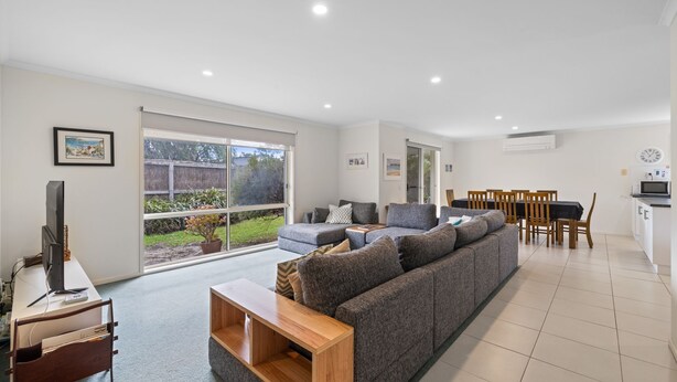 A modern living room with a grey sectional sofa, a wooden coffee table, and a TV on a white stand. There is a dining area with wooden chairs and a table, and a kitchen area with a microwave and a clock on the wall. - Ohana Beach House Inverloch (INVERLOCH)
