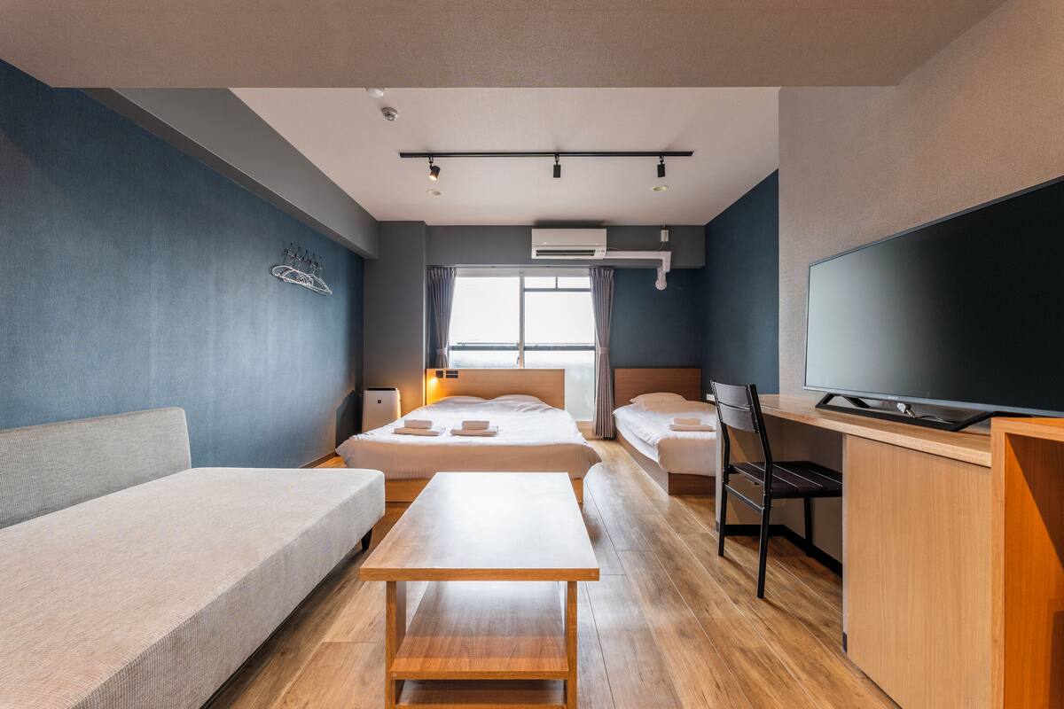 Una habitación de hotel moderna con sofá, dos camas individuales, un escritorio, televisor y un ventanal grande.