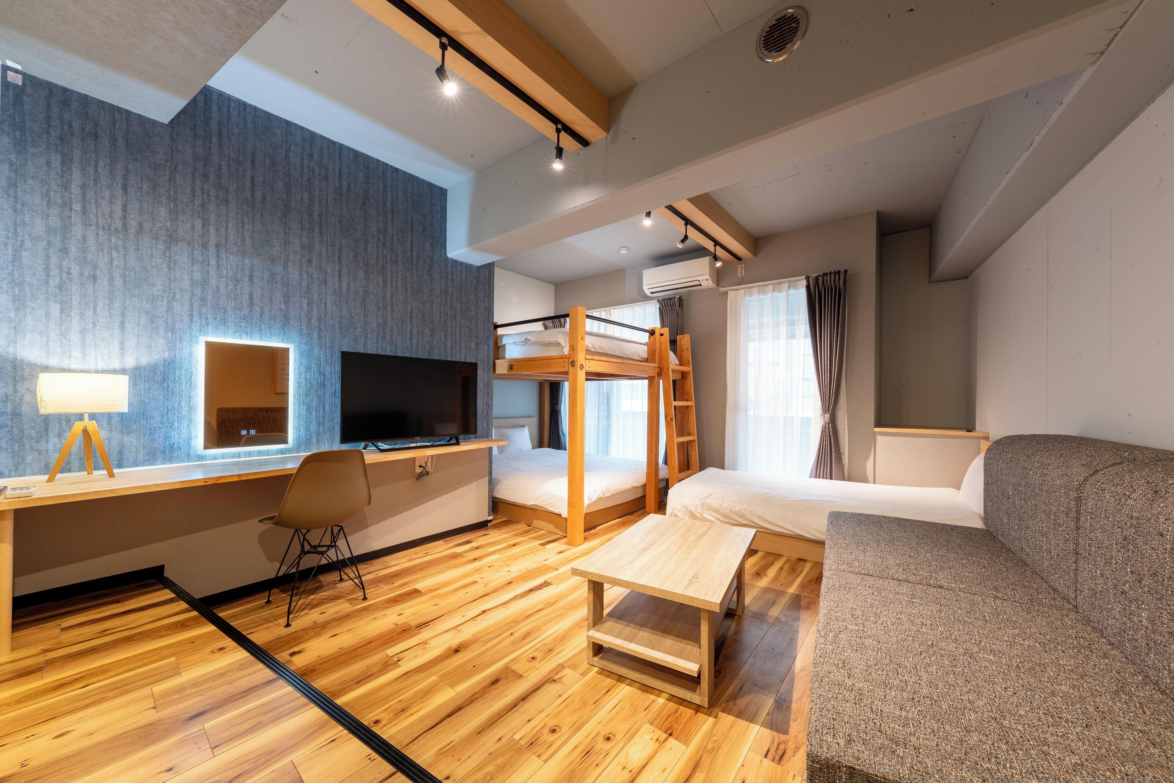 A modern hotel room with a bunk bed, a desk with a computer, a sofa, and a wooden coffee table.