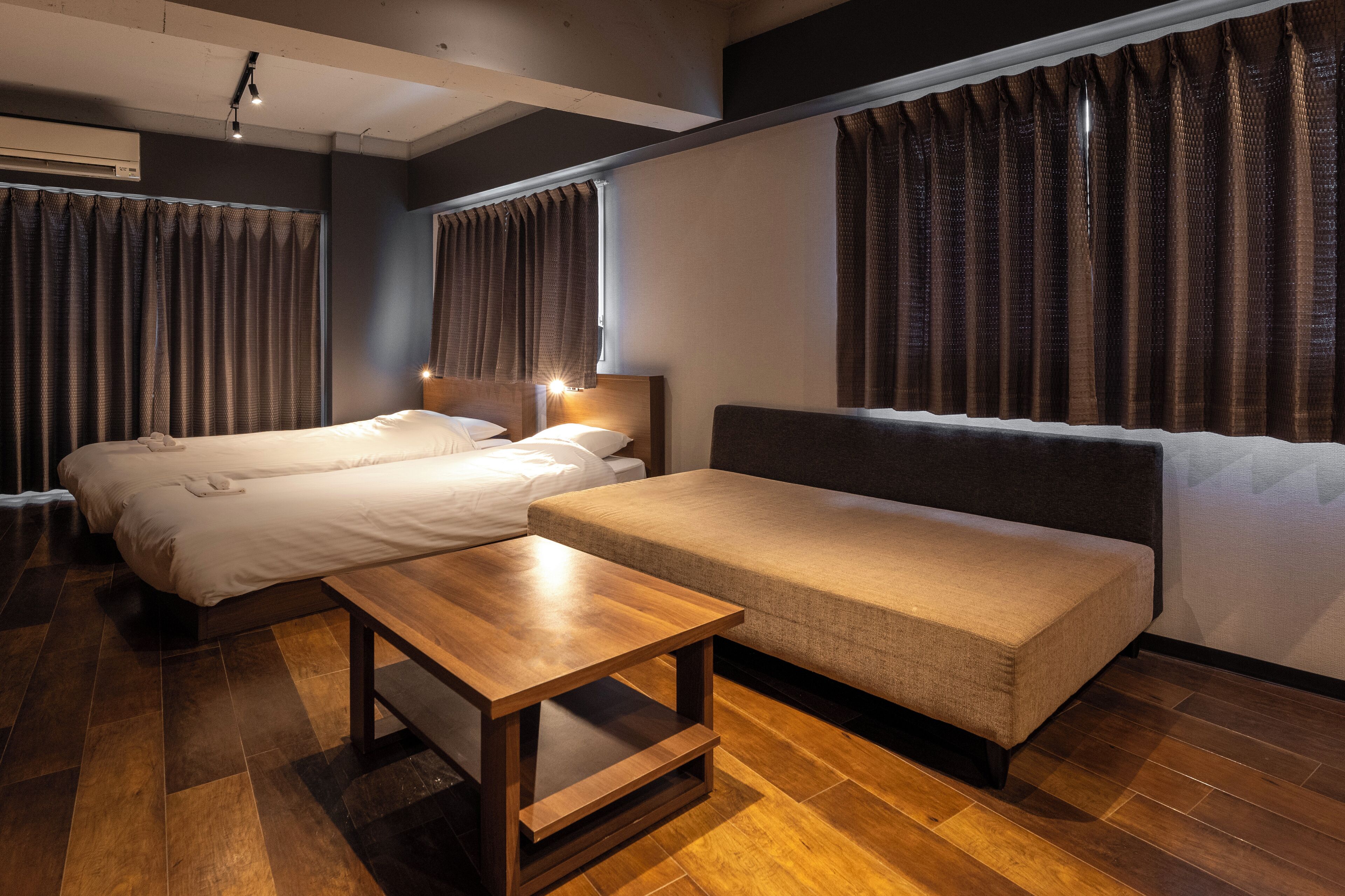 A hotel room with two beds, a sofa, a wooden table, and a large window with curtains.