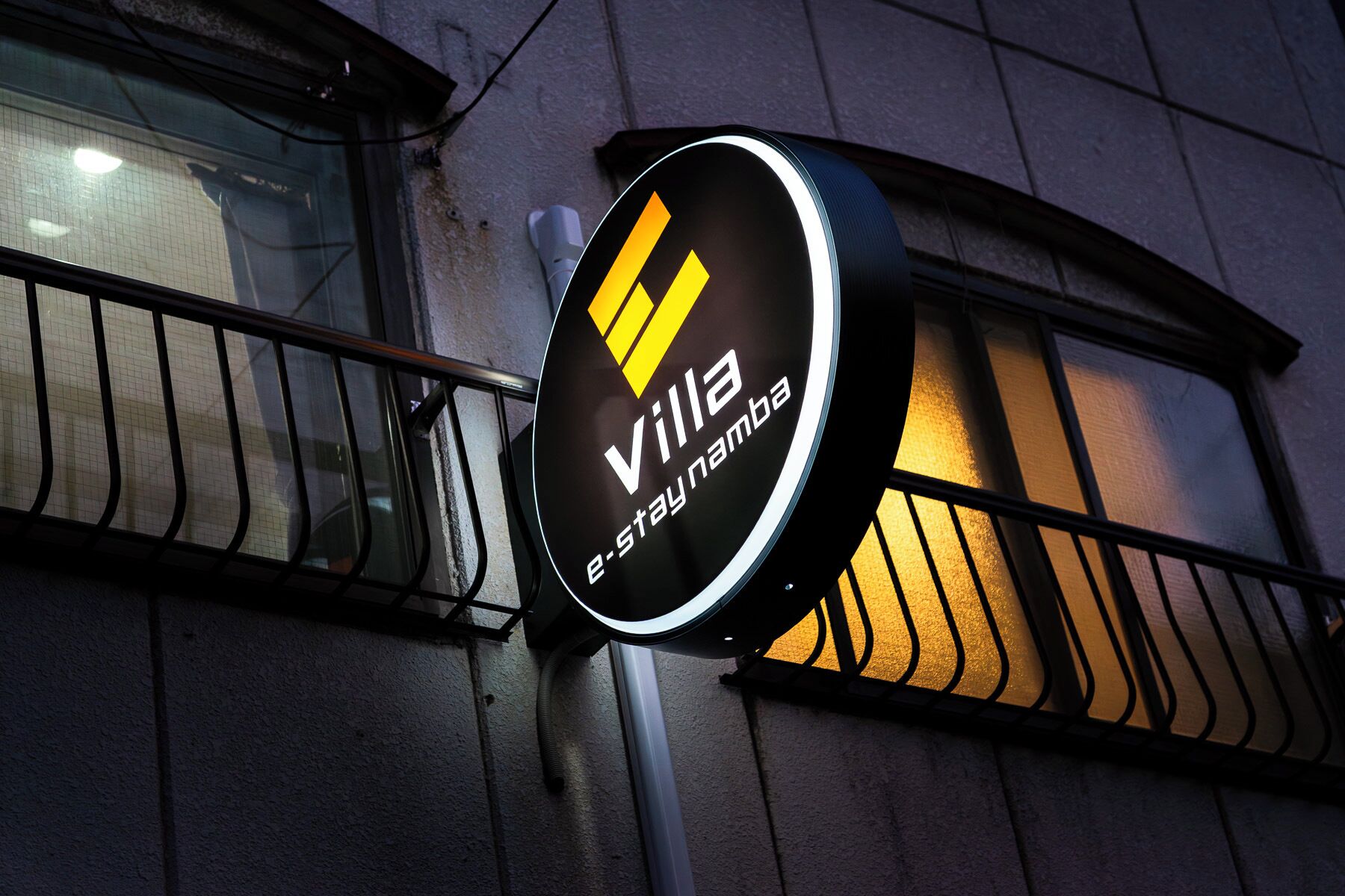 Un panneau circulaire portant l’inscription « villa e-stay namba » est fixé sur un poteau.