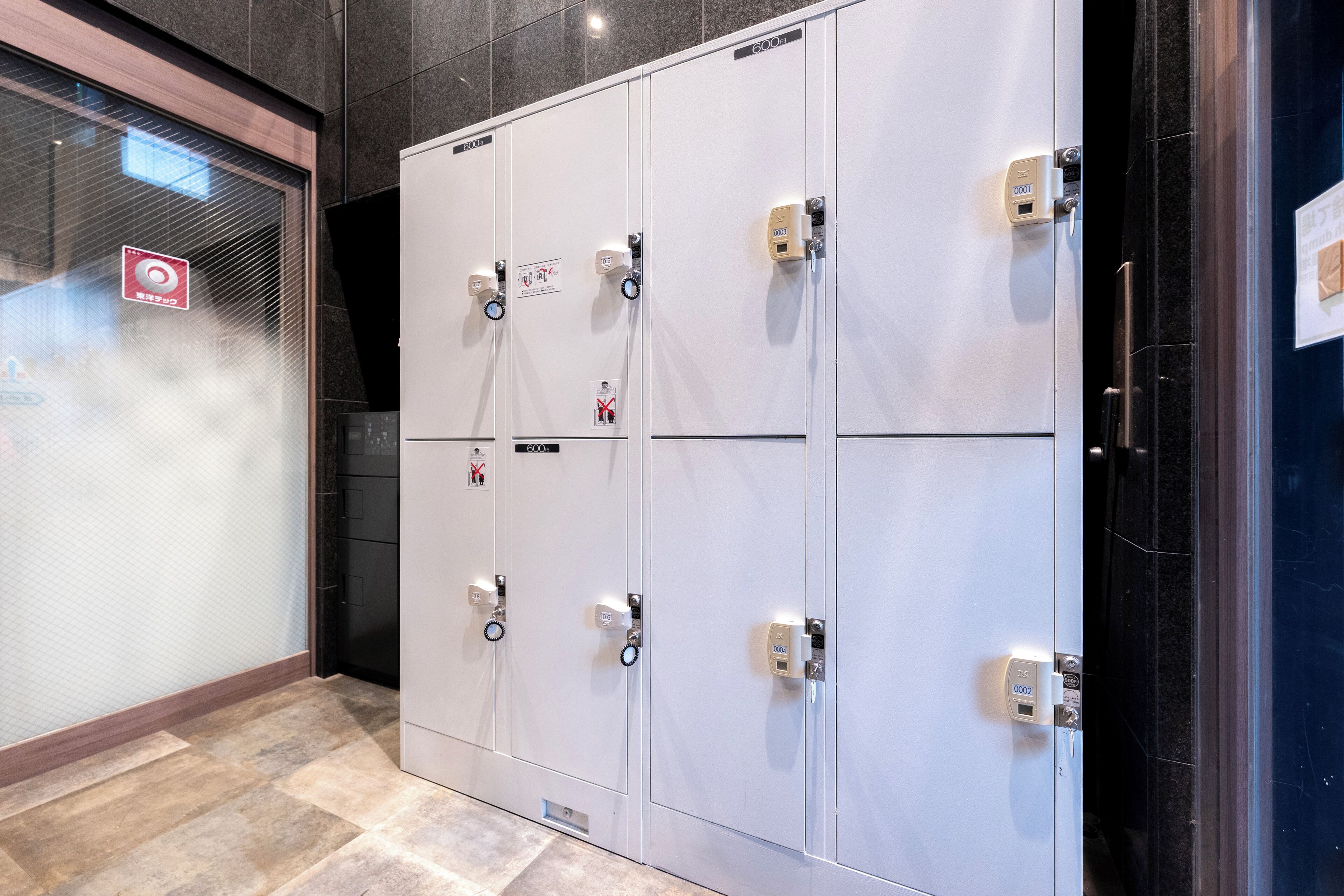 A row of white lockers with combination locks and a red emergency exit sign on the left.
