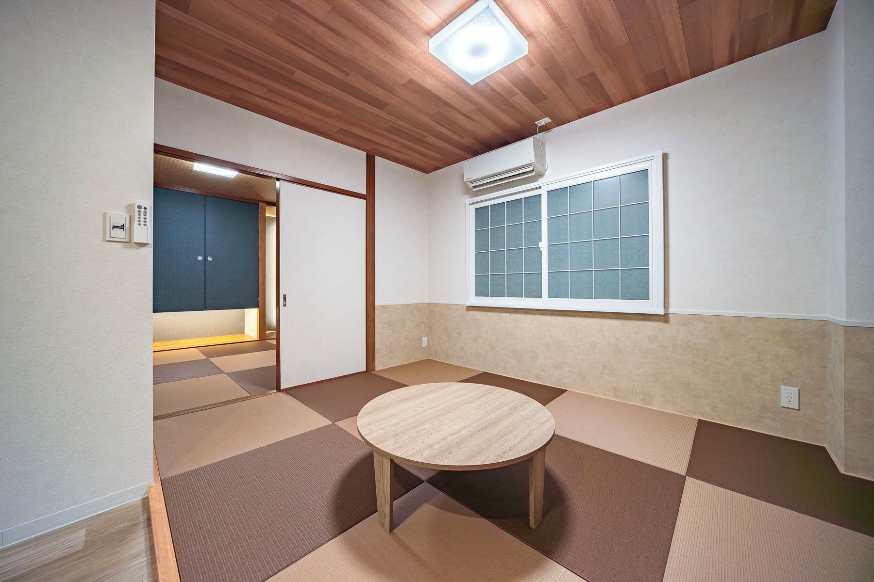 A room with tatami flooring, a wooden ceiling, a window with a grid pattern, and a small round table.