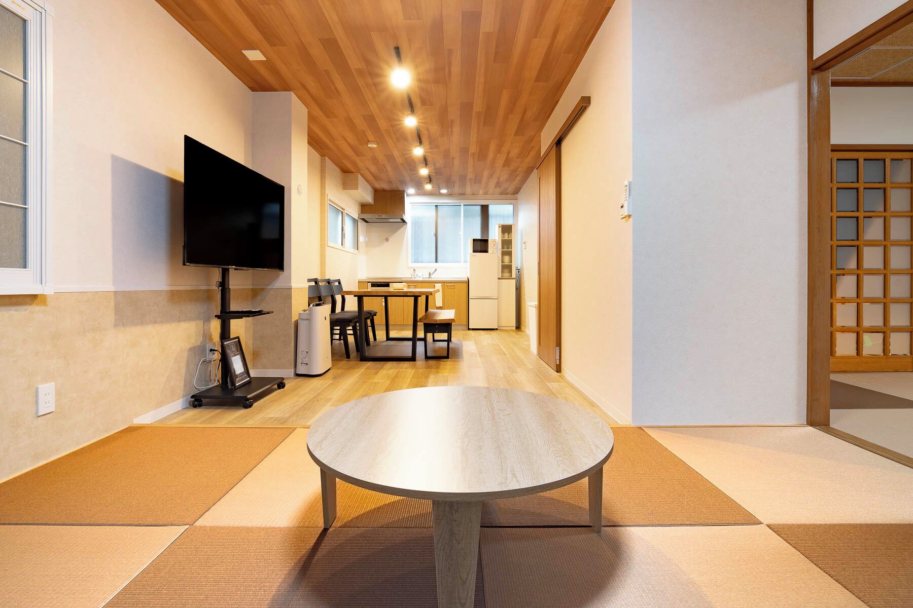 Une chambre d’hôtel moderne avec un plafond en bois, un téléviseur à écran plat fixé au mur, un coin repas avec une table et des chaises, et une cuisine en arrière-plan.