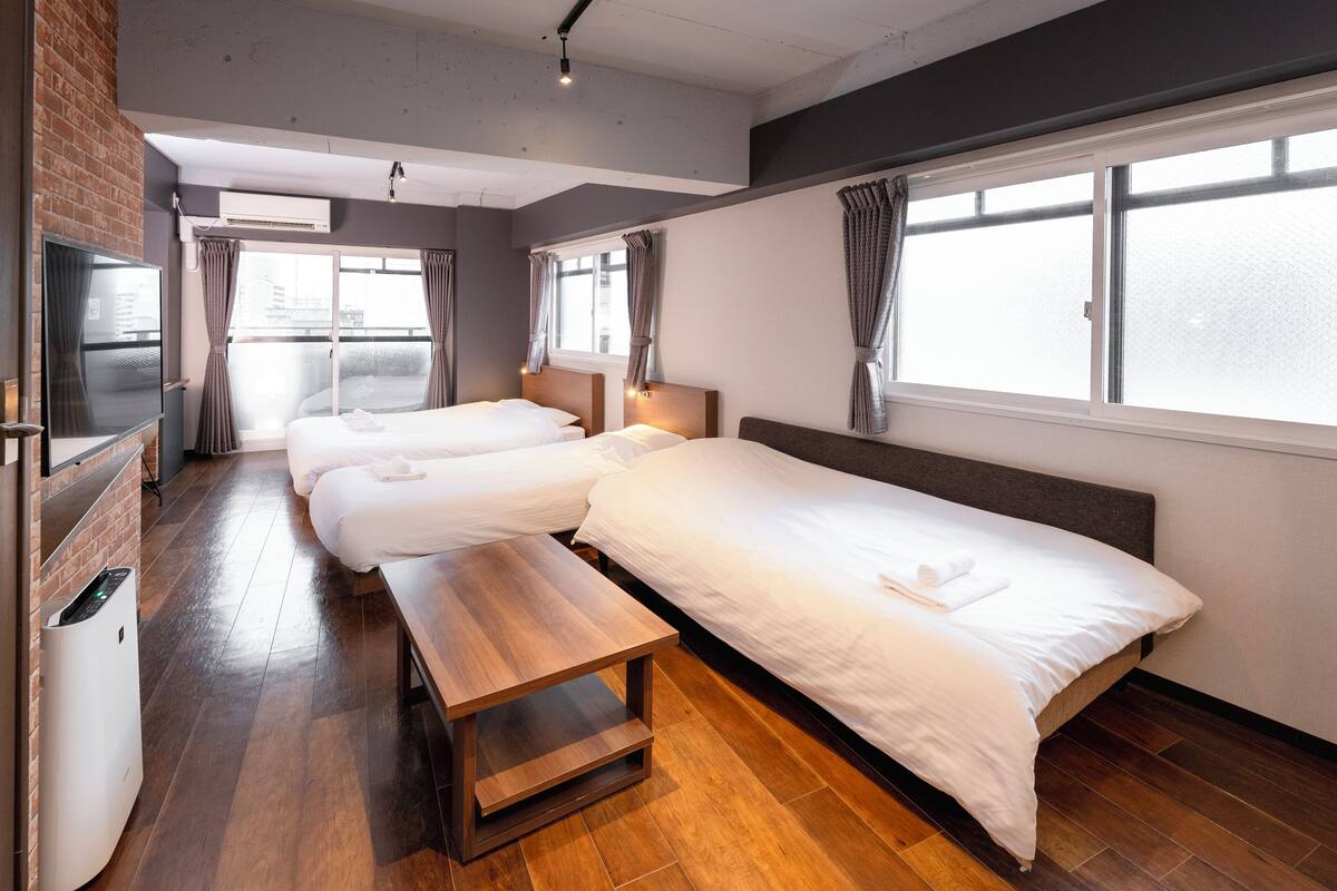 Una habitación de hotel con tres camas, un banco de madera, un televisor y una pared de ladrillo.
