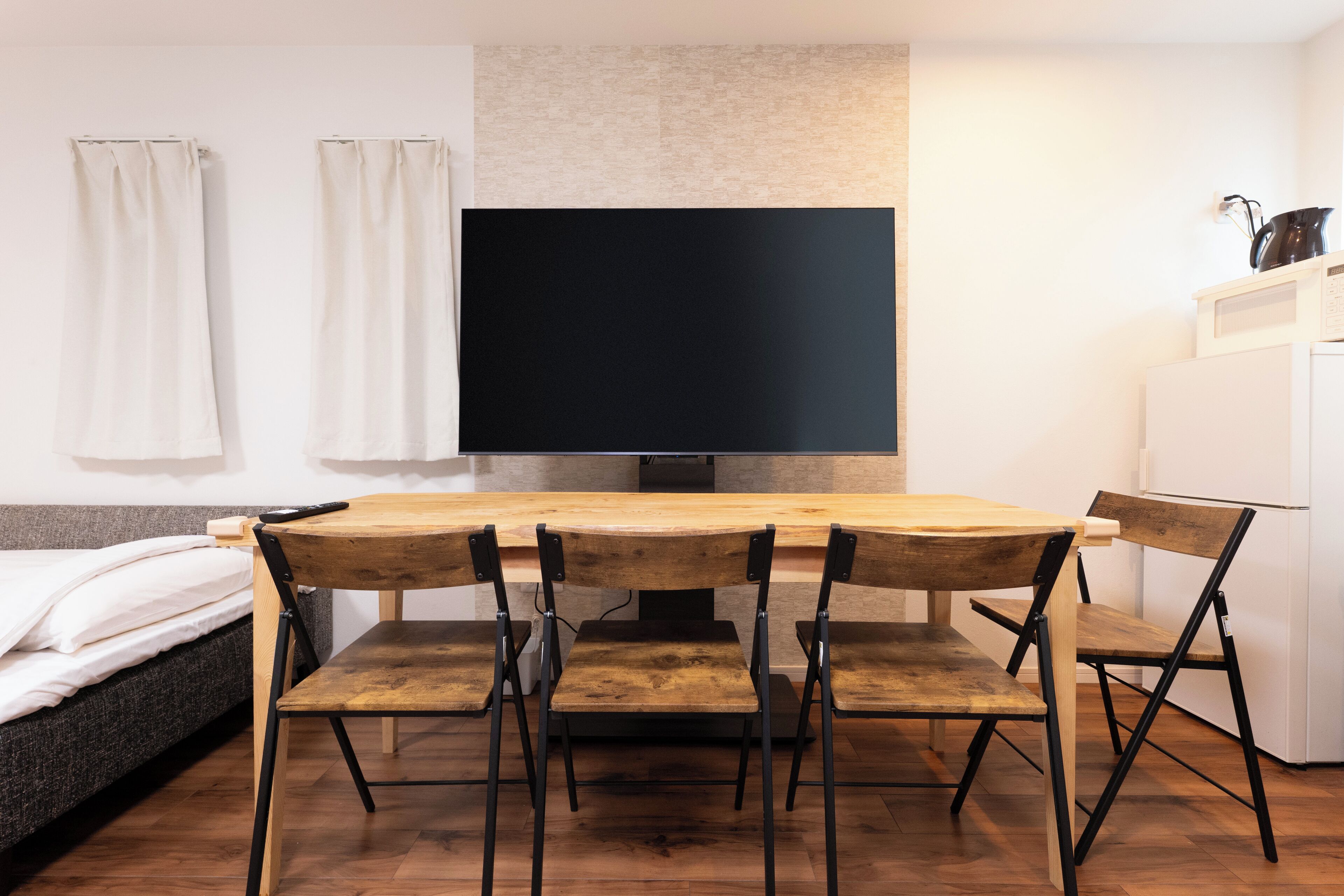 Ein kompakter Wohnraum mit Esstisch aus Holz, vier Stühlen, einem Flachbildfernseher, einem Bett mit weißen Leinen und einem weißen Kühlschrank.