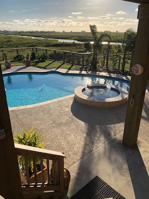 A swimming pool with a hot tub, surrounded by a concrete deck and a view of a river and greenery.
