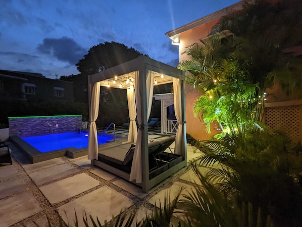 Un espace piscine extérieur privatif doté d’un coin salon couvert, d’un jacuzzi et d’une pergola illuminée de guirlandes lumineuses.