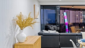 A modern living room with a white vase of dried plants on a wooden side table, a black sofa, and a glass partition with a view of a cityscape. - Circle on Cavill 3 Bedroom Ocean View — Q Stay (Surfers Paradise)