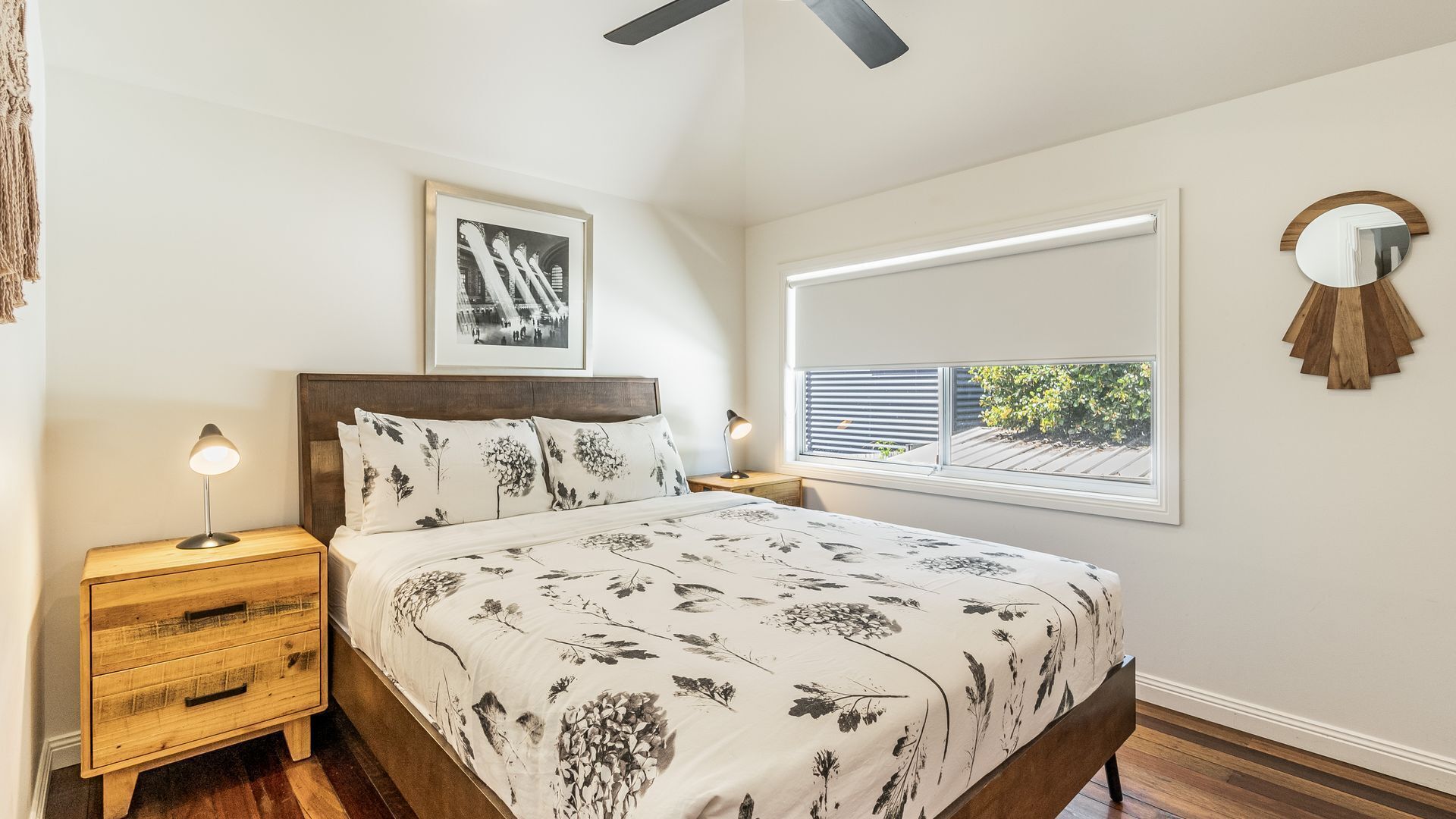 Un dormitorio con cama, mesitas de noche de madera, ventilador de techo, un cuadro enmarcado, una ventana con persianas y un espejo redondo.