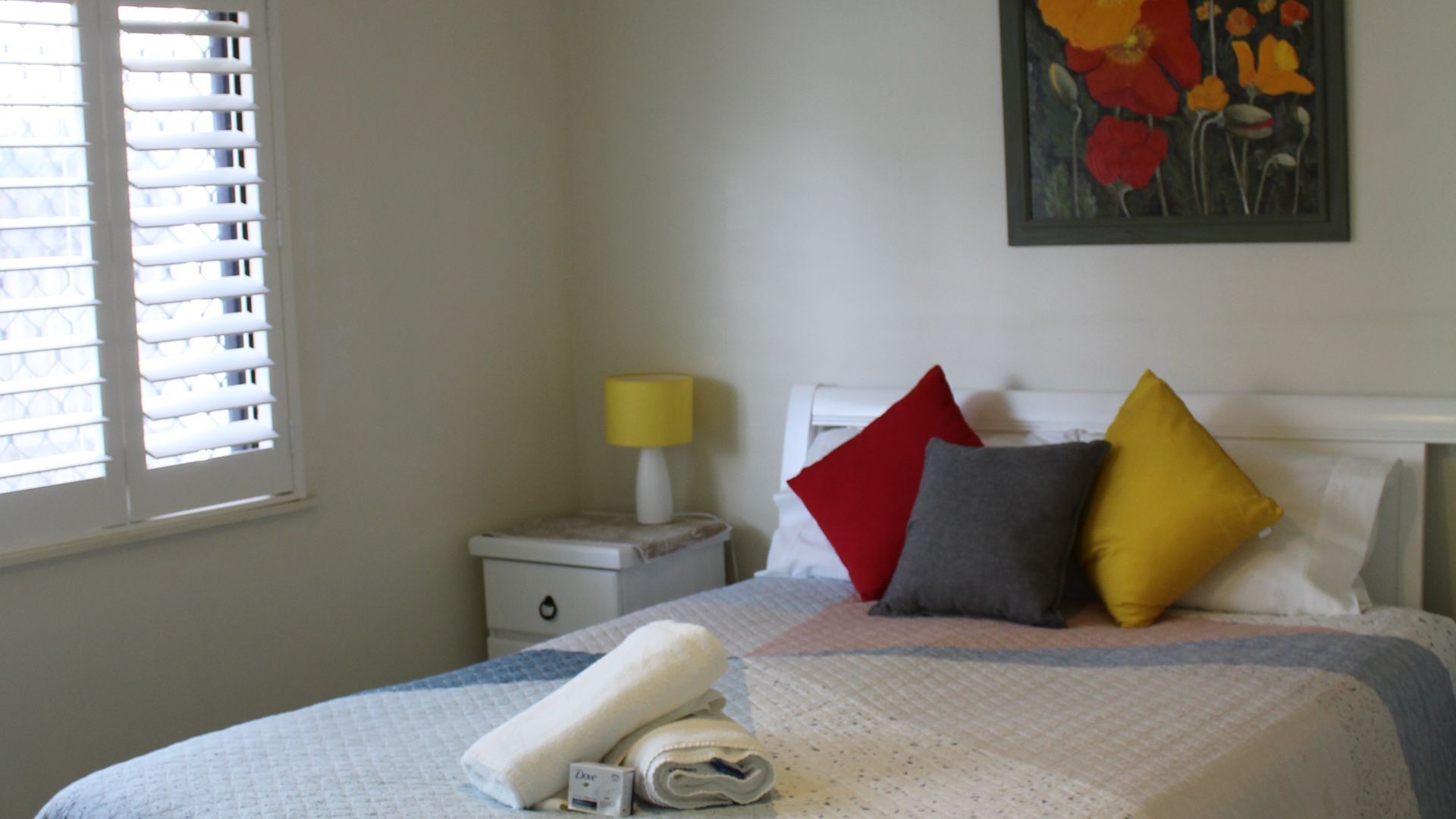 A neatly made bed with a white comforter, two pillows in red and yellow, and a rolled towel on the side.