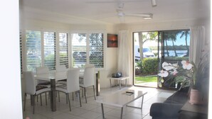A bright living room with a white sofa, a coffee table, and a dining area with white chairs and a table. There is a sliding glass door leading to a balcony with a view of the street and parked cars. - Noosaville River Front  - Watch the colors over the water (Noosaville)