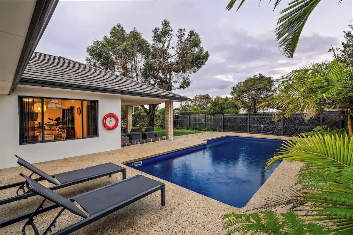 A swimming pool with lounge chairs, a covered patio, and a view of trees and a fence.