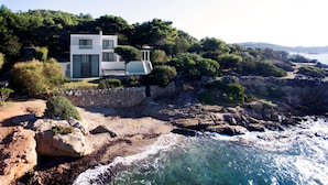Una casa moderna de dos pisos con un muro de contención de piedra, ubicada en una costa rocosa con vista al mar.