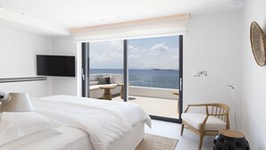 Un dormitorio moderno con una cama grande, un televisor de pantalla plana, una silla de madera y vistas al mar a través de amplios ventanales.