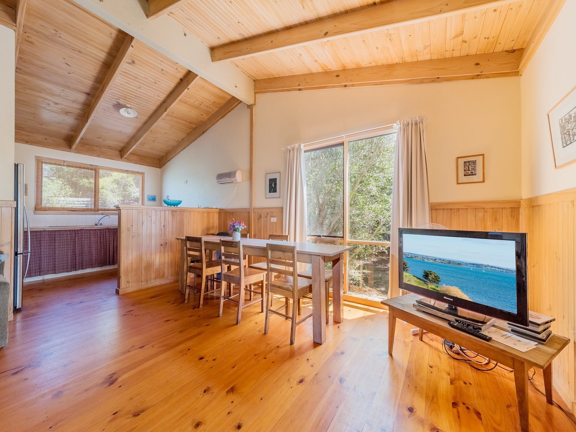 A cozy cabin interior with a wooden dining table, chairs, a television, and a large window overlooking a scenic view.