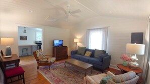A living room with a sofa, armchairs, a coffee table, and a TV. There is a kitchen in the background with a window and a ceiling fan.