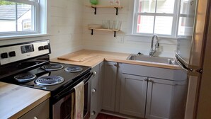 A compact kitchen with a stove, sink, and refrigerator. There are wooden shelves above the counter and a window providing natural light.
