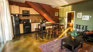 A modern kitchen with a central island, a dining area with stools, a staircase, and a living room with a coffee table and chairs. - Crooked Tree Guest Suites - The Fir Suite (Penticton)